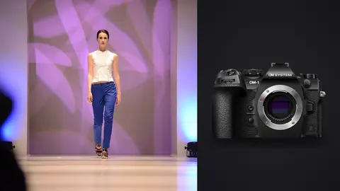 OM System OM-1 Mark II product photo beside model in a white top and blue trousers walking a runway