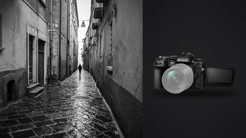 OM System OM-1 Mark II product photo beside lone figure walking a narrow rain-slicked old-town alley