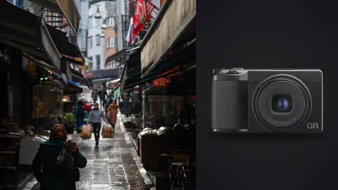 Ricoh GR IIIx product photo beside shopper walking a narrow rain-slicked market alley