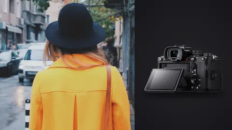 Sony Alpha 1 product photo beside woman in a mustard jacket and black hat walks a city street