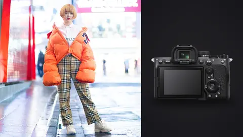 Sony a7 IV product photo beside woman in an orange puffer jacket and plaid trousers poses on a street
