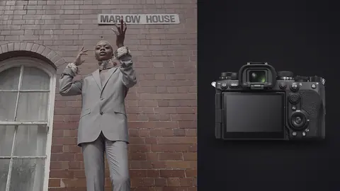 Sony a9 III product photo beside person in a gray suit gestures against a brick wall near a sign