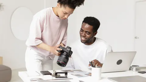 Two photographers reviewing images on a camera together at a desk with prints