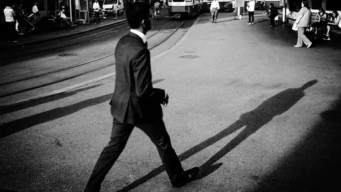 A man in a suit strides across a city tram square, casting a long shadow
