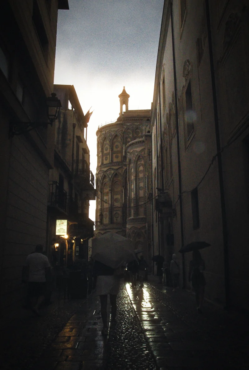 Une ruelle étroite à Monreale, en Sicile, un soir de pluie : des passants avec des parapluies sur des pavés mouillés, l’enseigne éclairée d’un bar à vin, la cathédrale au bout de la rue